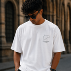 Person wearing a white t-shirt with a logo and text, standing in front of a stone building.